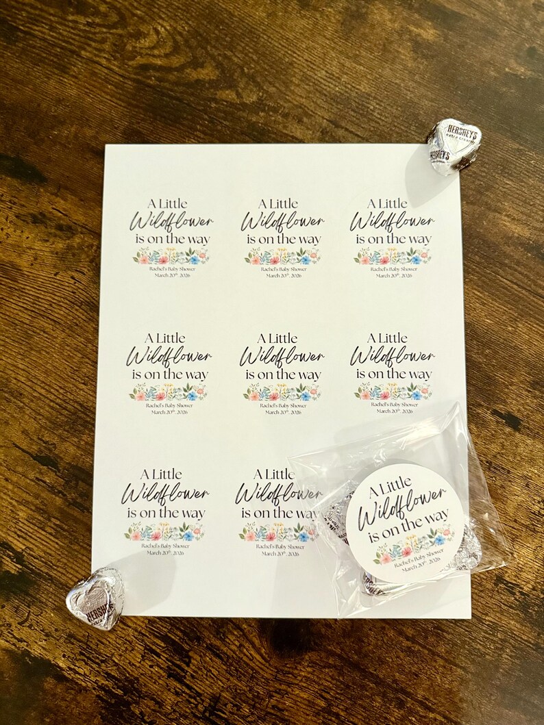 May include: A sheet of stickers and a round sticker with the text "A Little Wildflower is on the way" and floral designs. The stickers are for a baby shower. Two Hershey's Kisses are on the wooden surface.