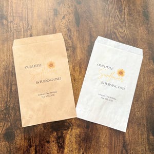 Puede incluir: Dos bolsas de papel, una beige y otra blanca, con el texto "OUR LITTLE Sunflower IS TURNING ONE!" y un gr&aacute;fico de girasol. Las bolsas tambi&eacute;n incluyen el texto "Jessica's First Birthday May 10th, 2026".