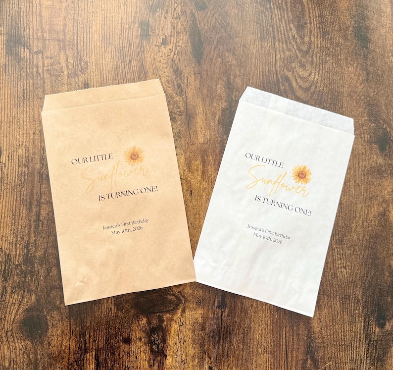 Puede incluir: Dos bolsas de papel, una beige y otra blanca, con el texto "OUR LITTLE Sunflower IS TURNING ONE!" y un gr&aacute;fico de girasol. Las bolsas tambi&eacute;n incluyen el texto "Jessica's First Birthday May 10th, 2026".