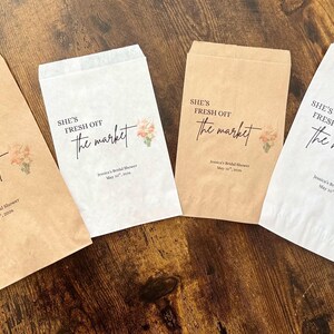 May include: Four paper favor bags in shades of brown and white, arranged on a wooden surface. Each bag features the text "She's Fresh Off the Market" in elegant script, along with a floral design and event details.