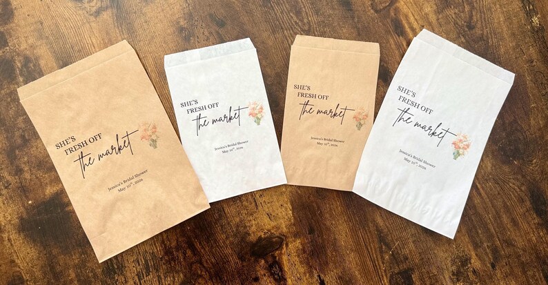 May include: Four paper favor bags in shades of brown and white, arranged on a wooden surface. Each bag features the text "She's Fresh Off the Market" in elegant script, along with a floral design and event details.