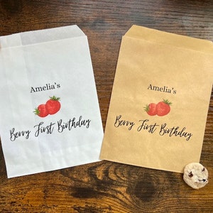 May include: Two paper treat bags, one white and one brown, with the text "Amelia's Berry First Birthday" and a strawberry graphic. A chocolate chip cookie rests near each bag. The bags are on a wooden surface.
