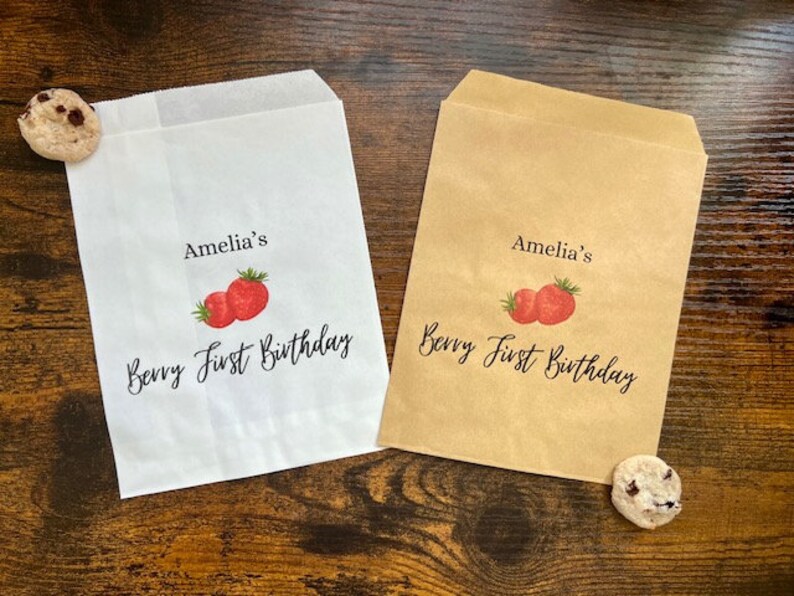 May include: Two paper treat bags, one white and one brown, with the text "Amelia's Berry First Birthday" and a strawberry graphic. A chocolate chip cookie rests near each bag. The bags are on a wooden surface.