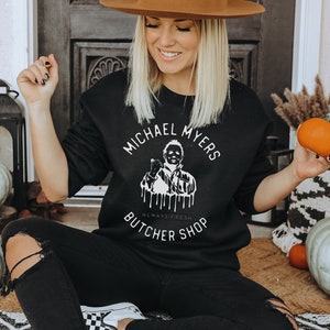 May include: Black sweatshirt with the text "MICHAEL MYERS BUTCHER SHOP" in a circular design. The graphic features a depiction of Michael Myers. The person in the image is wearing black pants and a brown hat. Pumpkins and gourds are in the background.