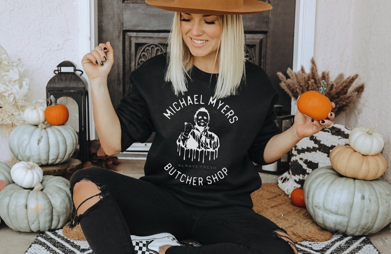 May include: Black sweatshirt with the text "MICHAEL MYERS BUTCHER SHOP" in a circular design. The graphic features a depiction of Michael Myers. The person in the image is wearing black pants and a brown hat. Pumpkins and gourds are in the background.
