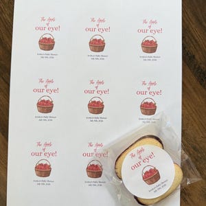 May include: A sheet of nine circular stickers featuring a watercolor illustration of a basket of red apples and the text The Apple of our eye! Jessica's Baby Shower July 15th, 2026. One sticker is applied to a cookie inside a clear plastic bag.
