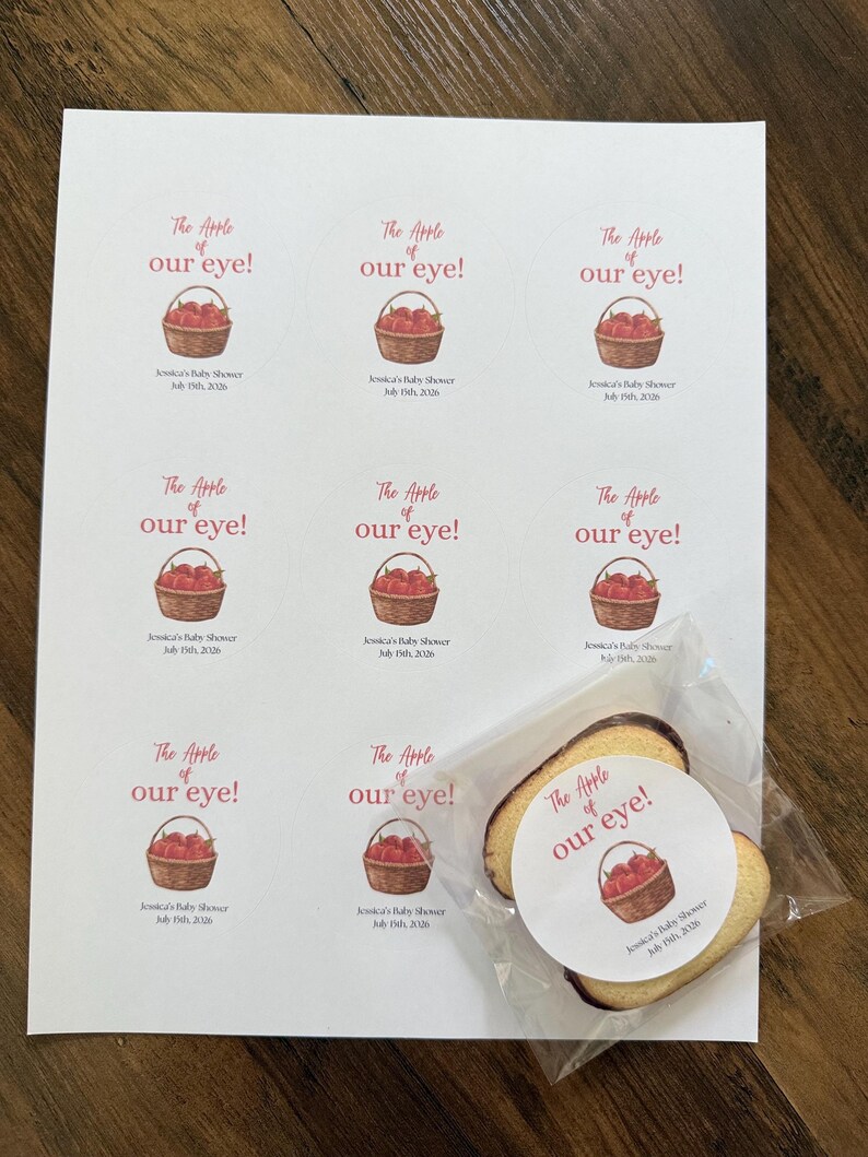 May include: A sheet of nine circular stickers featuring a watercolor illustration of a basket of red apples and the text The Apple of our eye! Jessica's Baby Shower July 15th, 2026. One sticker is applied to a cookie inside a clear plastic bag.