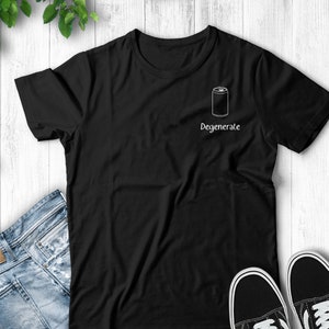 Op de afbeelding: Zwart T-shirt met een witte omtrek van een blikje en het woord "Degenerate". Het shirt wordt getoond met een blauwe jeans en zwart-witte sneakers op een witte houten achtergrond.