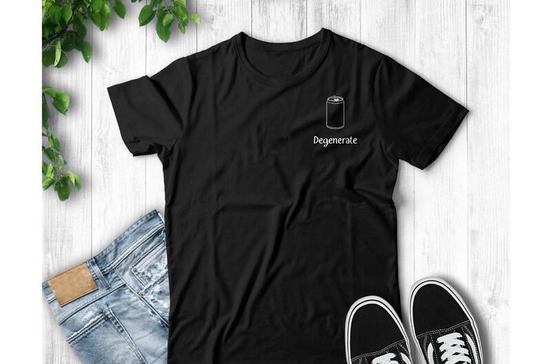 Op de afbeelding: Zwart T-shirt met een witte omtrek van een blikje en het woord "Degenerate". Het shirt wordt getoond met een blauwe jeans en zwart-witte sneakers op een witte houten achtergrond.