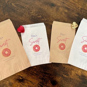 May include: Four paper treat bags with the words "Two Sweet" in red script, a donut graphic, and text. Two bags are brown, and two are white. Each bag is decorated with a heart-shaped candy.