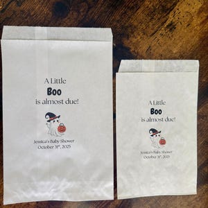 K&ouml;nnte beinhalten: Zwei wei&szlig;e Papiert&uuml;ten mit dem Text "A Little Boo is almost due!" und einer Geisterillustration, die einen K&uuml;rbis h&auml;lt. Die T&uuml;ten sind f&uuml;r Jessicas Babyparty am 31. Oktober 2025.