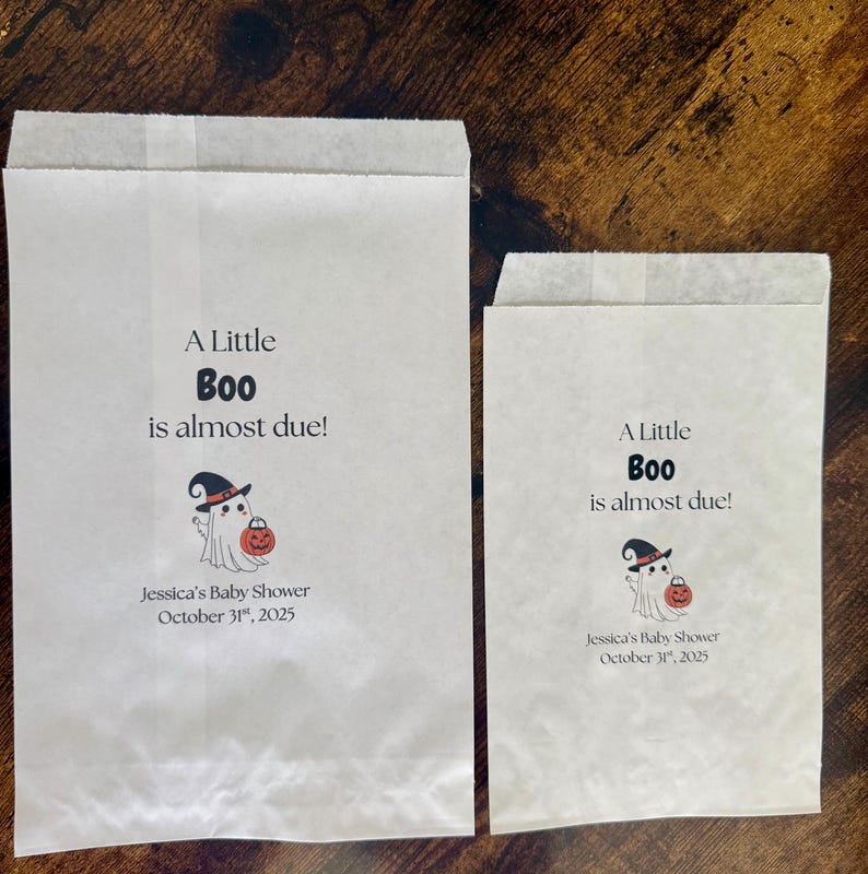 K&ouml;nnte beinhalten: Zwei wei&szlig;e Papiert&uuml;ten mit dem Text "A Little Boo is almost due!" und einer Geisterillustration, die einen K&uuml;rbis h&auml;lt. Die T&uuml;ten sind f&uuml;r Jessicas Babyparty am 31. Oktober 2025.