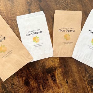 Puede incluir: Cuatro bolsas de papel para regalos, dos en marr&oacute;n kraft y dos en blanco, est&aacute;n dispuestas sobre una superficie de madera. Cada bolsa presenta el texto "She Found Her Main Squeeze" con una ilustraci&oacute;n de naranja y hojas.
