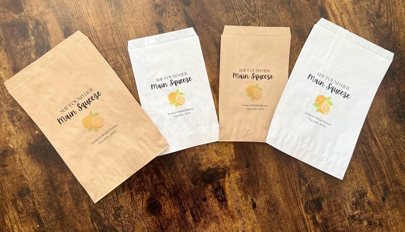 Puede incluir: Cuatro bolsas de papel para regalos, dos en marr&oacute;n kraft y dos en blanco, est&aacute;n dispuestas sobre una superficie de madera. Cada bolsa presenta el texto "She Found Her Main Squeeze" con una ilustraci&oacute;n de naranja y hojas.