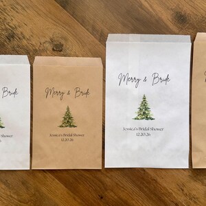 May include: Four rectangular paper favor bags in white and kraft brown colors, each featuring a printed green Christmas tree, the text Merry & Bride, Jessica's Bridal Shower, and the date 12.20.26, accompanied by a small round chocolate treat.
