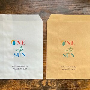 May include: Two paper favor bags, one white and one brown, with the text "ONE in the SUN" in colorful letters. Below the text is "Nick's First Birthday August 17th, 2024".