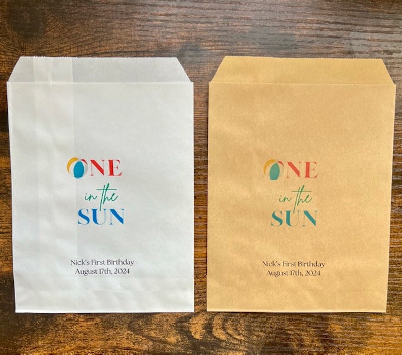 May include: Two paper favor bags, one white and one brown, with the text "ONE in the SUN" in colorful letters. Below the text is "Nick's First Birthday August 17th, 2024".
