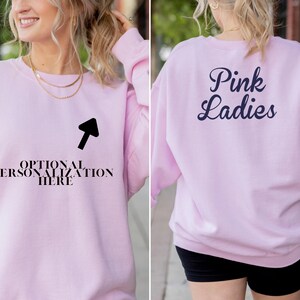May include: A light pink sweatshirt with the text "Pink Ladies" on the back in navy blue script. The front of the sweatshirt has the text "OPTIONAL PERSONALIZATION HERE" with an arrow pointing to it. The sweatshirt is a crew neck style.