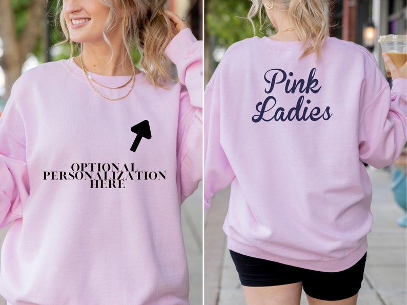 May include: A light pink sweatshirt with the text "Pink Ladies" on the back in navy blue script. The front of the sweatshirt has the text "OPTIONAL PERSONALIZATION HERE" with an arrow pointing to it. The sweatshirt is a crew neck style.