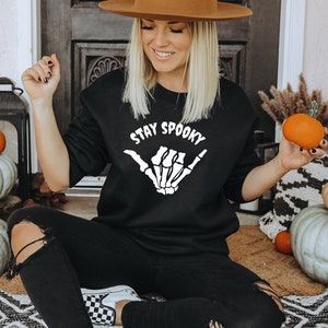 Może przedstawiać: Czarna bluza z napisem "STAY SPOOKY" i białym wzorem szkieletu dłoni. Osoba trzyma małą pomarańczową dynię. Tematem jest Halloween.