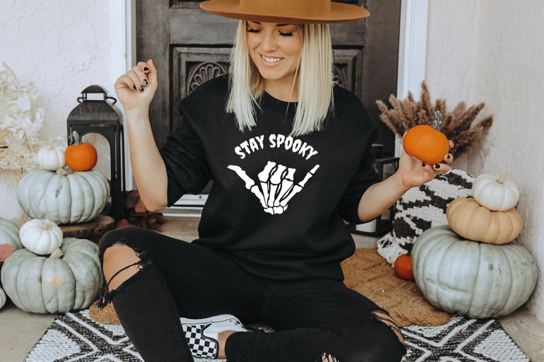 Może przedstawiać: Czarna bluza z napisem "STAY SPOOKY" i białym wzorem szkieletu dłoni. Osoba trzyma małą pomarańczową dynię. Tematem jest Halloween.