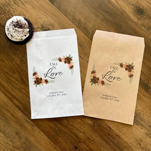 Puede incluir: Dos bolsas de papel rectangulares, una blanca y otra marr&oacute;n, con un gr&aacute;fico de corona floral, el texto "FALL IN Love" y los nombres "Jessica & Dave" con la fecha "October 30th, 2026". Una galleta oscura con glaseado blanco est&aacute; al lado.