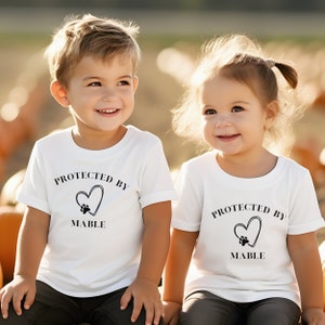 May include: Two children wearing white t-shirts with the text "PROTECTED BY MABLE" above a heart and paw print design. The children are smiling, and the background is blurred with pumpkins.