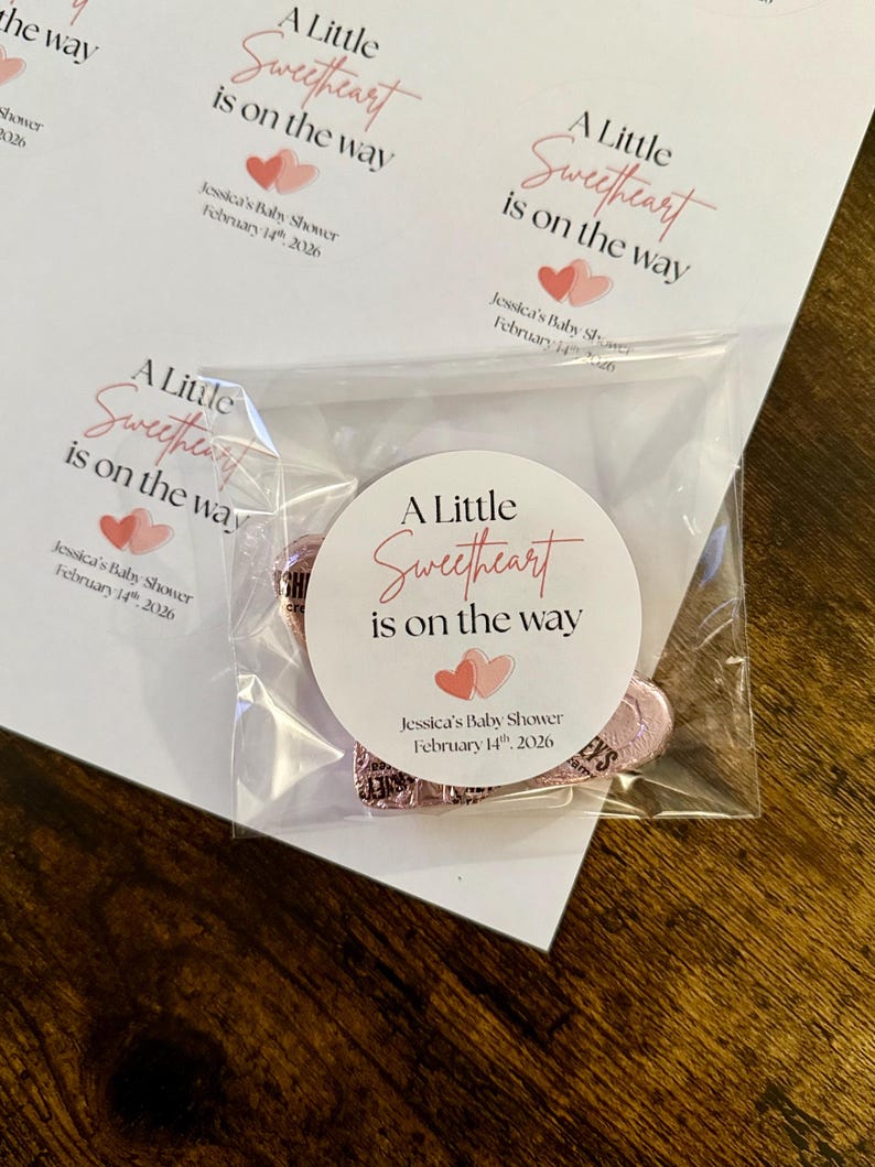 May include: A cellophane bag with a round sticker and pink sweets. The sticker says "A Little Sweetheart is on the way" with a baby shower date. The background shows printed sheets with the same text and design.