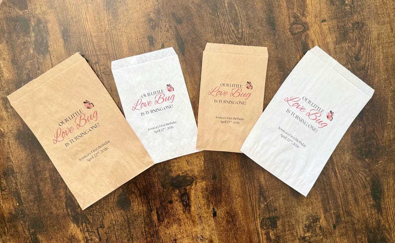 May include: Four paper gift bags, two brown and two white, are arranged on a wooden surface. Each bag has the text "Our Little Love Bug is Turning One!" printed in red, along with a butterfly graphic and a name and date.