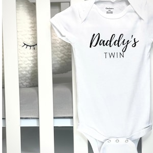 May include: White baby onesie with the text "Daddy's TWIN" in black script. The onesie is made of a soft material and has short sleeves. The onesie is hanging on a white crib.