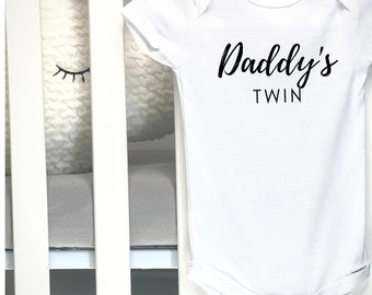 May include: White baby onesie with the text "Daddy's TWIN" in black script. The onesie is made of a soft material and has short sleeves. The onesie is hanging on a white crib.