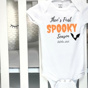 May include: White baby onesie with the text "Theo's First Spooky Season October 2023" in black and orange. A black bat graphic is on the right side. The onesie is made by Gerber and is size 3-6M.