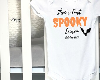May include: White baby onesie with the text "Theo's First Spooky Season October 2023" in black and orange. A black bat graphic is on the right side. The onesie is made by Gerber and is size 3-6M.