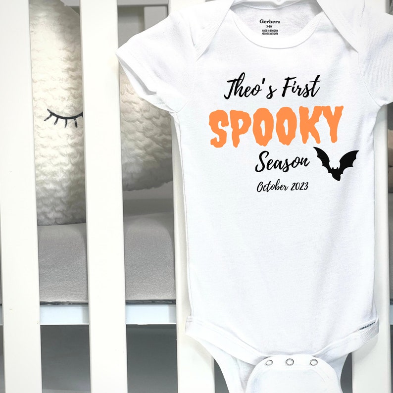 Puede incluir: Body de beb&eacute; blanco con el texto "Theo's First Spooky Season October 2023" en negro y naranja. Un gr&aacute;fico de murci&eacute;lago negro est&aacute; a la derecha. El body es de Gerber, talla 3-6M.