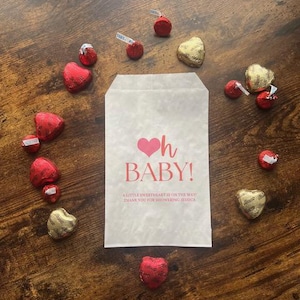 May include: A white paper bag with the text "oh BABY!" in pink, surrounded by heart-shaped and round chocolates in red and gold foil. The bag includes the message "A LITTLE SWEETHEART IS ON THE WAY! THANK YOU FOR SHOWERING JESSICA."