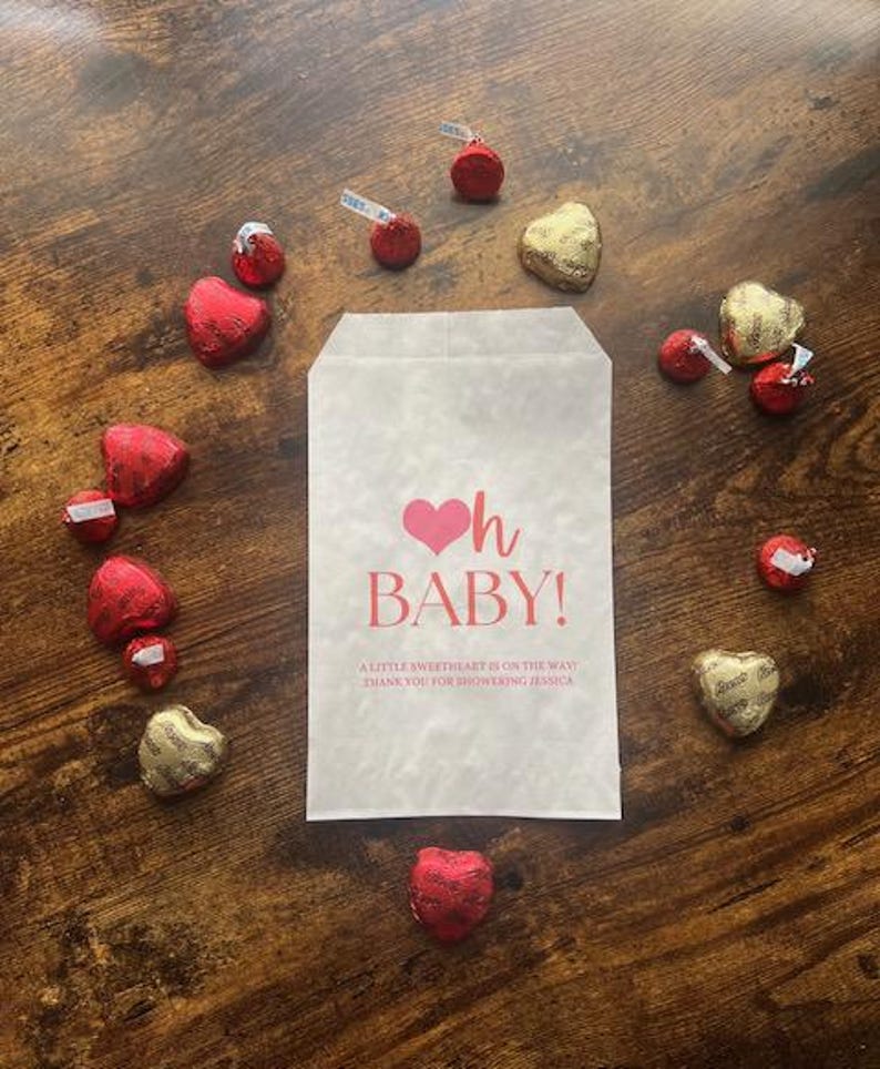 May include: A white paper bag with the text "oh BABY!" in pink, surrounded by heart-shaped and round chocolates in red and gold foil. The bag includes the message "A LITTLE SWEETHEART IS ON THE WAY! THANK YOU FOR SHOWERING JESSICA."