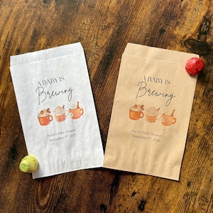 May include: Two paper treat bags, one white and one tan, with the text "A BABY IS Brewing" in elegant script. Below are illustrations of three mugs of coffee. The text "Kayla's Baby Shower November 5th, 2025" is printed below the mugs.