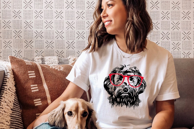 Puede incluir: Camiseta blanca con una ilustraci&oacute;n en blanco y negro de un perro con gafas rojas. La cara del perro es el punto central, con detalles de pelaje y las gafas que a&ntilde;aden un toque de color. La camiseta la lleva una persona.