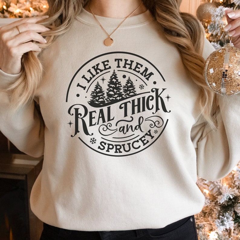 Pu&ograve; includere: Felpa beige con grafica nera che recita "I LIKE THEM REAL THICK and SPRUCEY" in un design circolare con pini e fiocchi di neve. La felpa &egrave; di colore neutro e ha un girocollo.