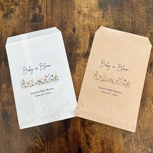 以下が含まれることがあります： 「Baby in Bloom」の文字が筆記体で書かれた、白とベージュの2つの紙袋。文字の下には花の模様があります。袋の下部には「Jessica's Baby Shower June 28, 2025」と書かれています。