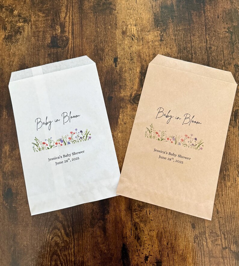 以下が含まれることがあります： 「Baby in Bloom」の文字が筆記体で書かれた、白とベージュの2つの紙袋。文字の下には花の模様があります。袋の下部には「Jessica's Baby Shower June 28, 2025」と書かれています。