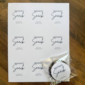 May include: A white sheet featuring eight circular stickers with the black text Midnight Snack, John & Jessica, and September 8th, 2026. One identical sticker is applied to a clear plastic bag containing a dark round cookie, placed over the sheet.