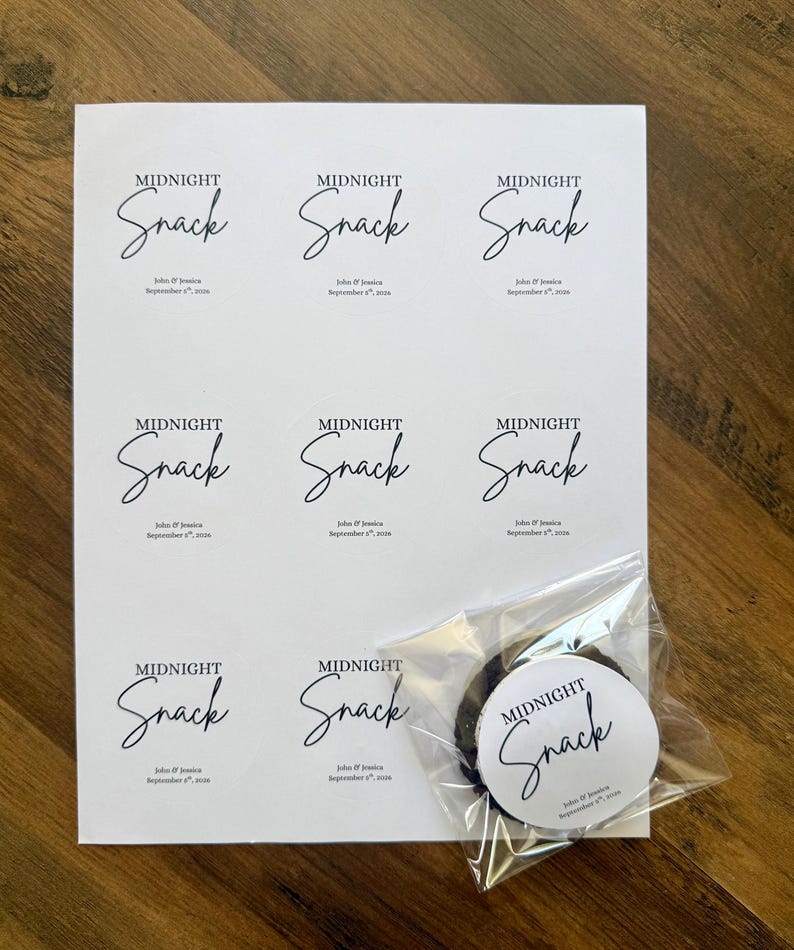 May include: A white sheet featuring eight circular stickers with the black text Midnight Snack, John & Jessica, and September 8th, 2026. One identical sticker is applied to a clear plastic bag containing a dark round cookie, placed over the sheet.