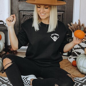 Może przedstawiać: Czarna bluza z napisem "Stay Spooky" i grafiką przedstawiającą szkielet dłoni. Osoba ma na sobie czarne dżinsy i brązowy kapelusz, trzymając małą pomarańczową dynię. Scena jest udekorowana dyniami i jesiennymi dekoracjami.