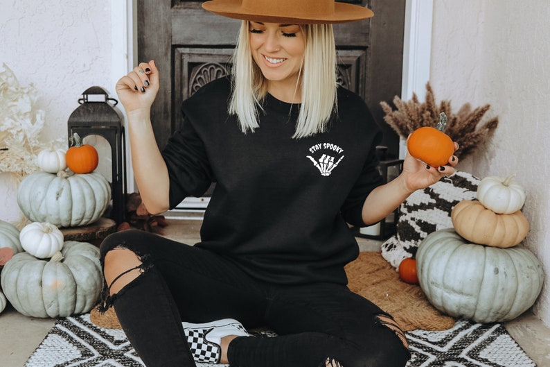 Może przedstawiać: Czarna bluza z napisem "Stay Spooky" i grafiką przedstawiającą szkielet dłoni. Osoba ma na sobie czarne dżinsy i brązowy kapelusz, trzymając małą pomarańczową dynię. Scena jest udekorowana dyniami i jesiennymi dekoracjami.