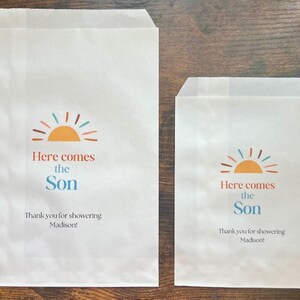 以下が含まれることがあります： カラフルな太陽のグラフィックと「Here comes the Son」の文字が入った2つの白い紙のトリートバッグ。バッグには「Thank you for showering Madison！」の文字も含まれています。チョコレートチップクッキーが各バッグの近くにあります。