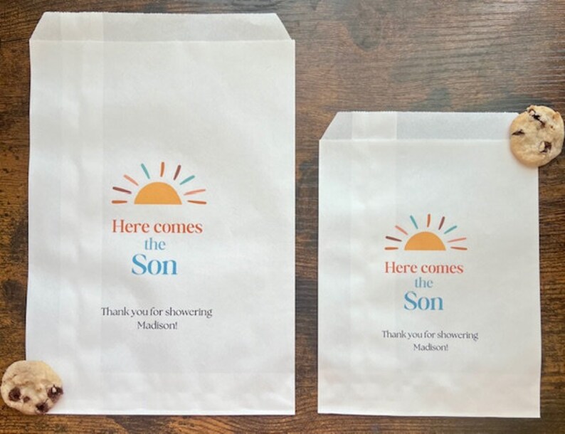 以下が含まれることがあります： カラフルな太陽のグラフィックと「Here comes the Son」の文字が入った2つの白い紙のトリートバッグ。バッグには「Thank you for showering Madison！」の文字も含まれています。チョコレートチップクッキーが各バッグの近くにあります。