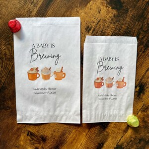 May include: Two white paper bags with the text "A Baby Is Brewing" and illustrations of coffee mugs. The bags also include the text "Kayla's Baby Shower November 5th, 2025". A red candy and a green candy are on the wooden surface.