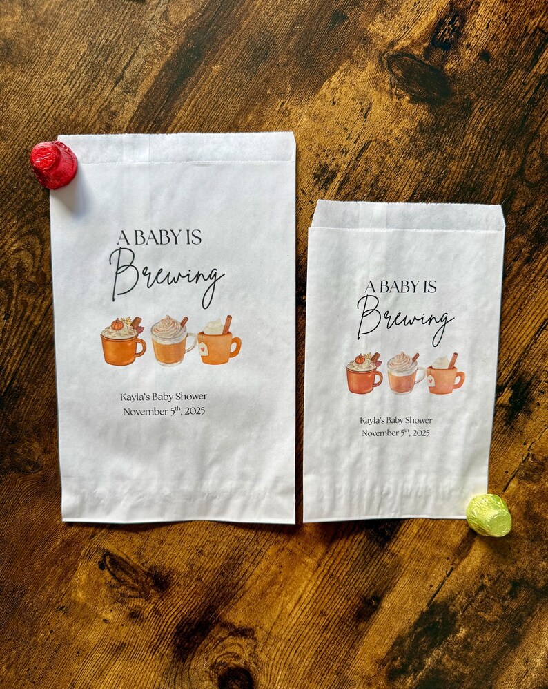 May include: Two white paper bags with the text "A Baby Is Brewing" and illustrations of coffee mugs. The bags also include the text "Kayla's Baby Shower November 5th, 2025". A red candy and a green candy are on the wooden surface.