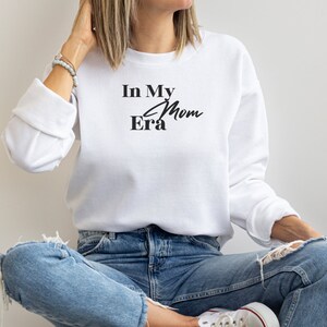 May include: White sweatshirt with the text "In My Mom Era" in black script. The long-sleeved top is paired with blue jeans. The model is wearing white sneakers.